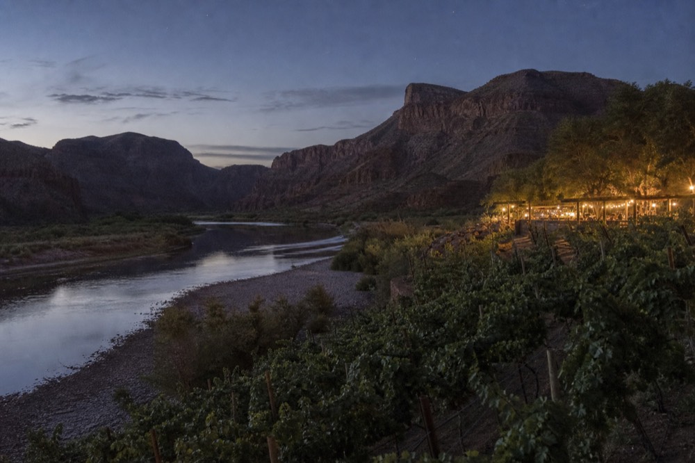 A wide bend of a desert river with a gravel beach on the near bank and planted vine rows rising on the terrace above it