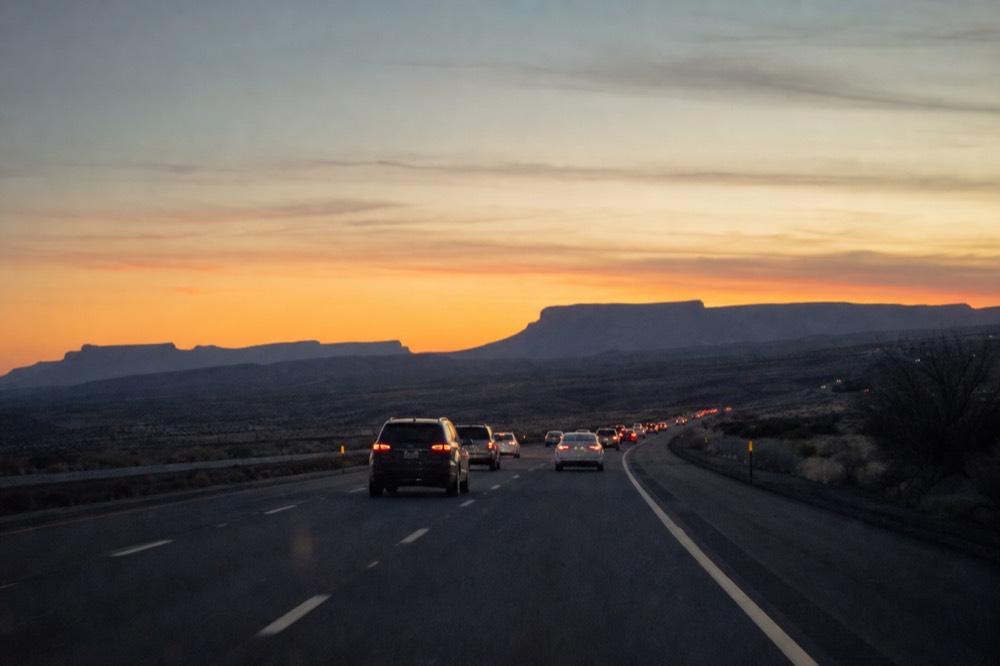 Wide desert interstate at dusk with taillights strung out ahead and mesas silhouetted against an orange sky