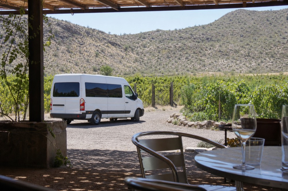 Small white shuttle van parked on a gravel drive beside a vineyard gate with hills rising behind it