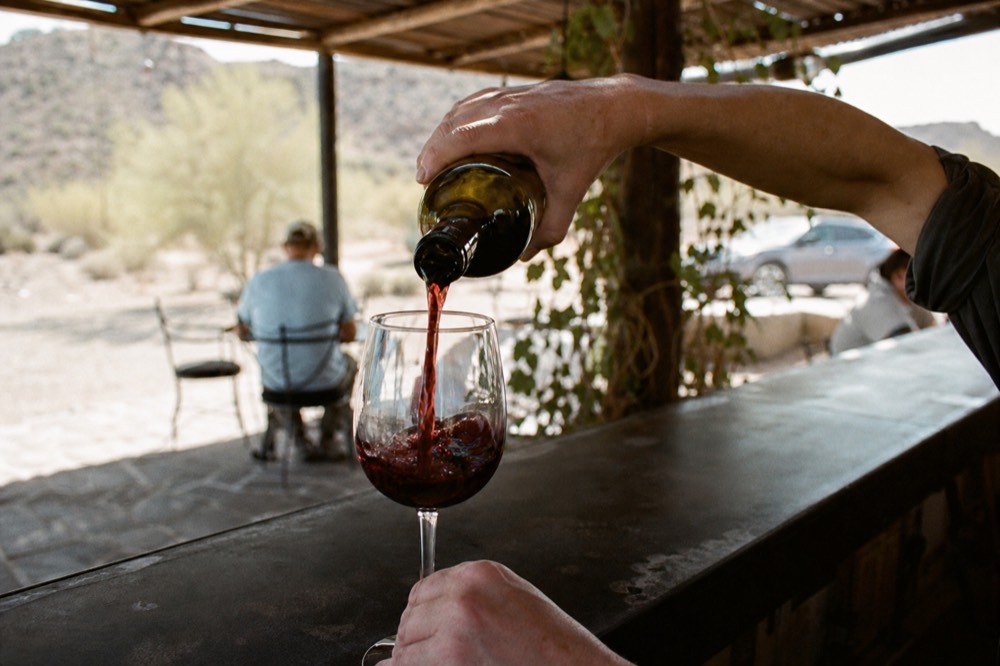 Hands pouring a dark red wine into a stemmed glass at an outdoor bar with vines blurred behind