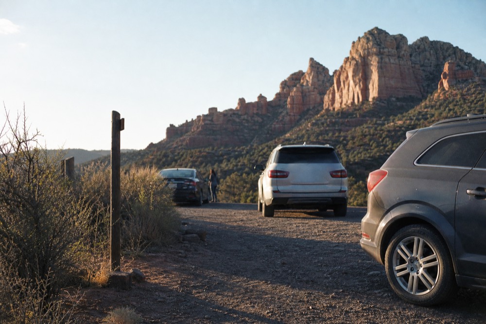 Small gravel parking pullout beside a desert road with a few cars and a wooden trail sign at first light