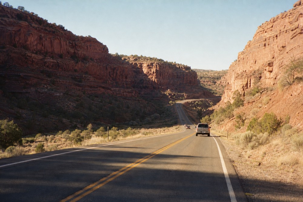Empty two-lane highway climbing between red sandstone cliffs in late afternoon light with long shadows across the asphalt