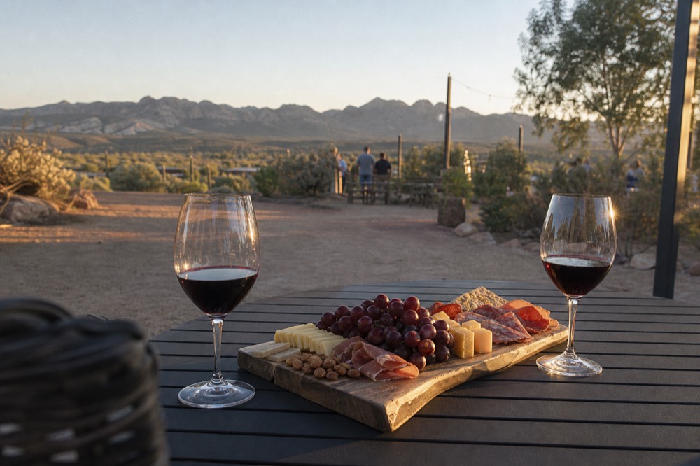 A wooden charcuterie board with cheese, cured meat and grapes beside two glasses of red wine on an outdoor table at golden hour