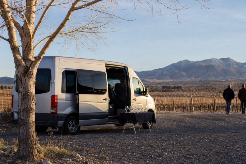 A small silver passenger van parked on gravel under a cottonwood tree beside a vineyard fence, side door open, folding table with glasses nearby