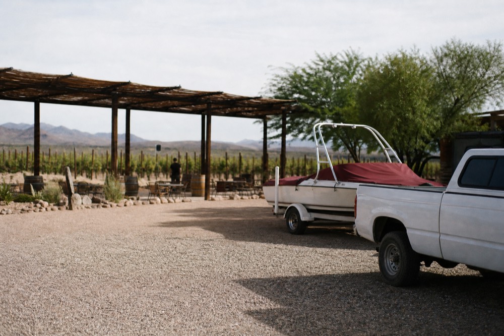 A gravel parking area beside a rural vineyard with a shade ramada, a boat trailer and vine rows stretching behind