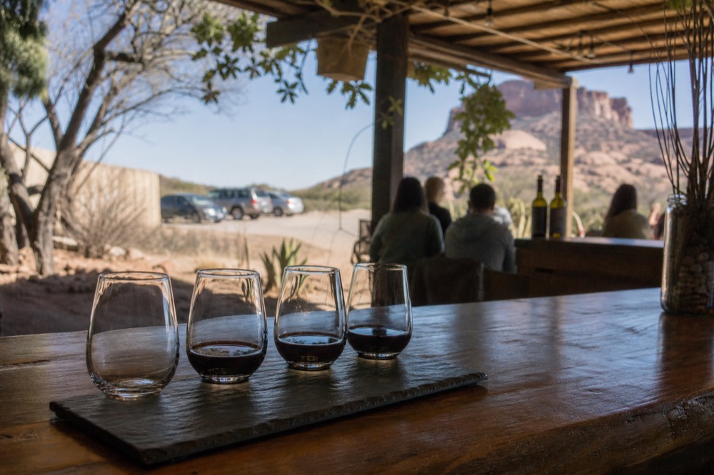 A wooden tasting counter with four small pour glasses lined up on a slate flight board, warm interior light, red rock visible through a window behind